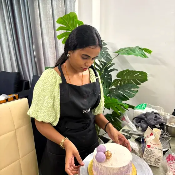 A student finishing a lilac cake with gold and purple spheres on a cake stand