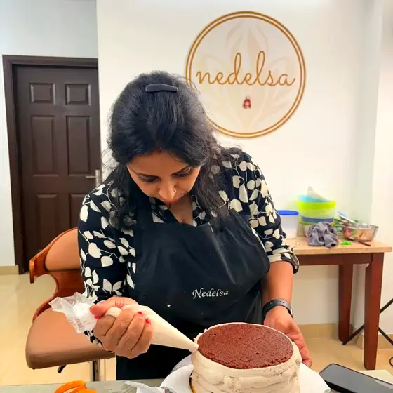 A student piping frosting onto a chocolate layer cake on a turntable in front of the Nedelsa logo wall